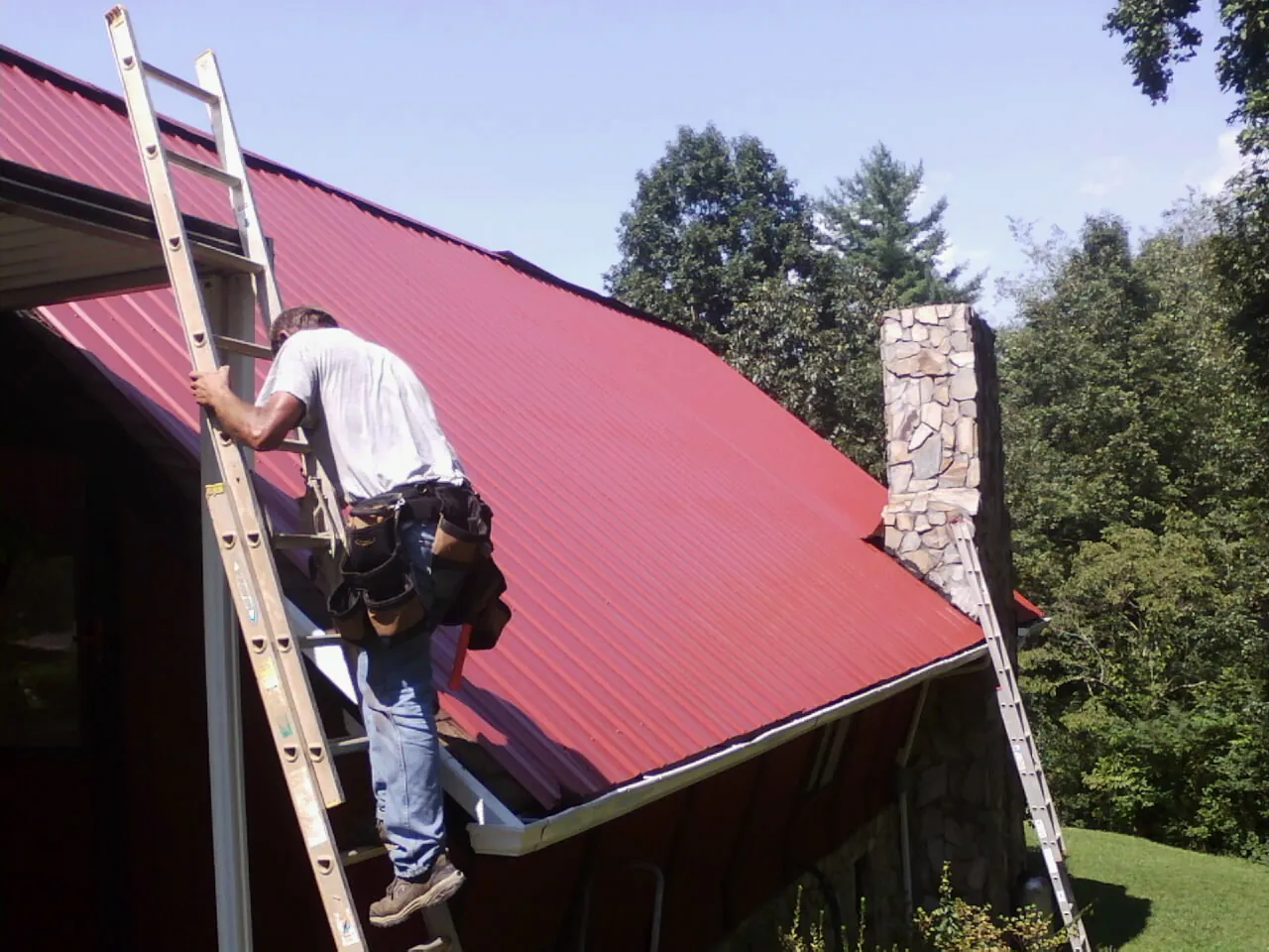 Roofer on a ladder beside a red metal roof and stone chimney — Soffit & Fascia Repair by Roofing Brothers in Lorain
